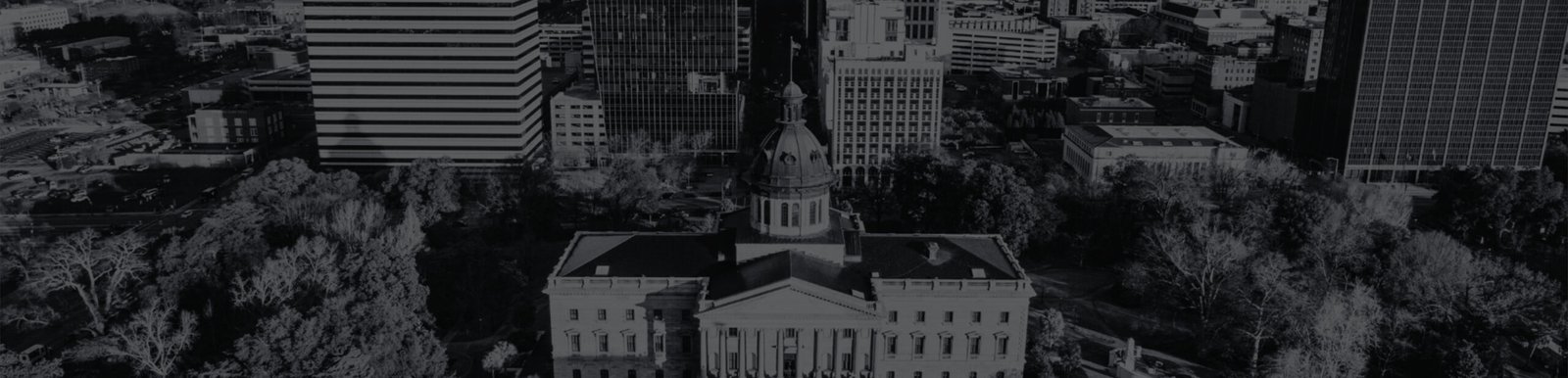 Aerial view of a government building with a dome, surrounded by trees and modern office buildings in a cityscape.