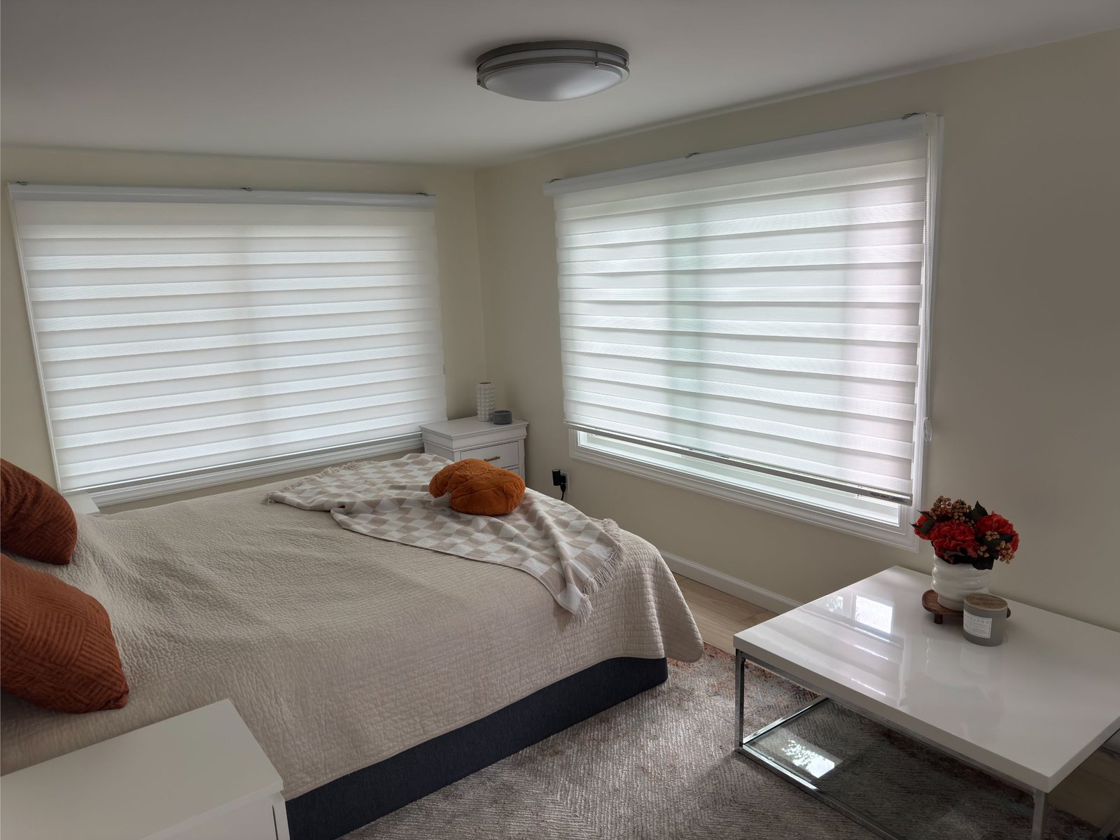 A neatly made bed with orange pillows, a blanket, and a teddy bear sits next to two large windows with white blinds in a bright, minimalist bedroom.