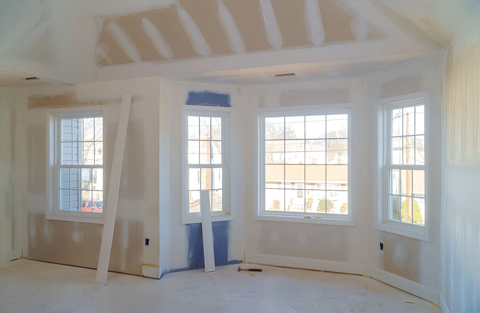 A room under construction with unfinished drywall, taped seams, three large windows, and materials on the floor.