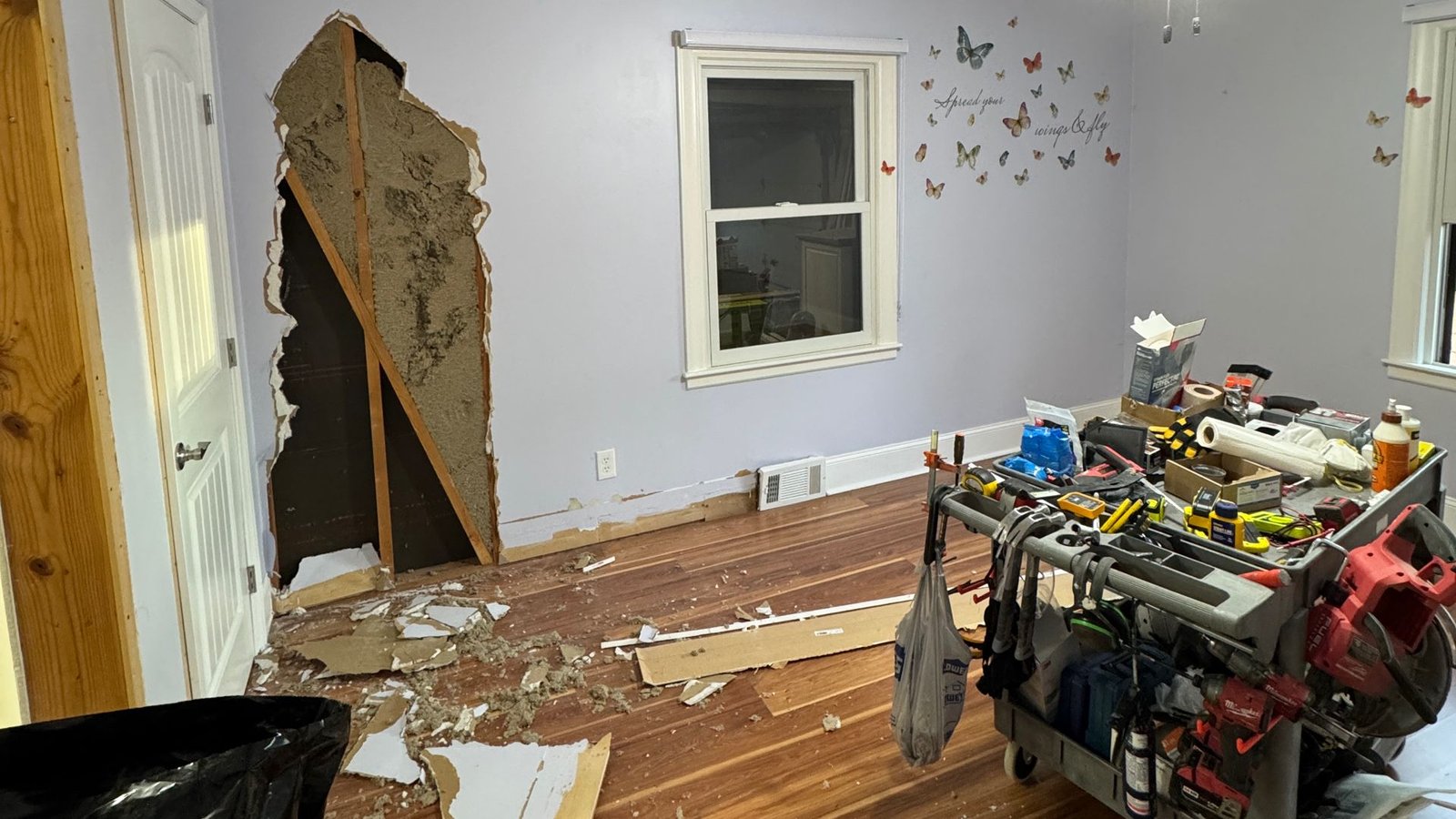 A room with a large hole in the wall, scattered drywall debris on the floor, and a tool cart with various construction tools and supplies nearby.
