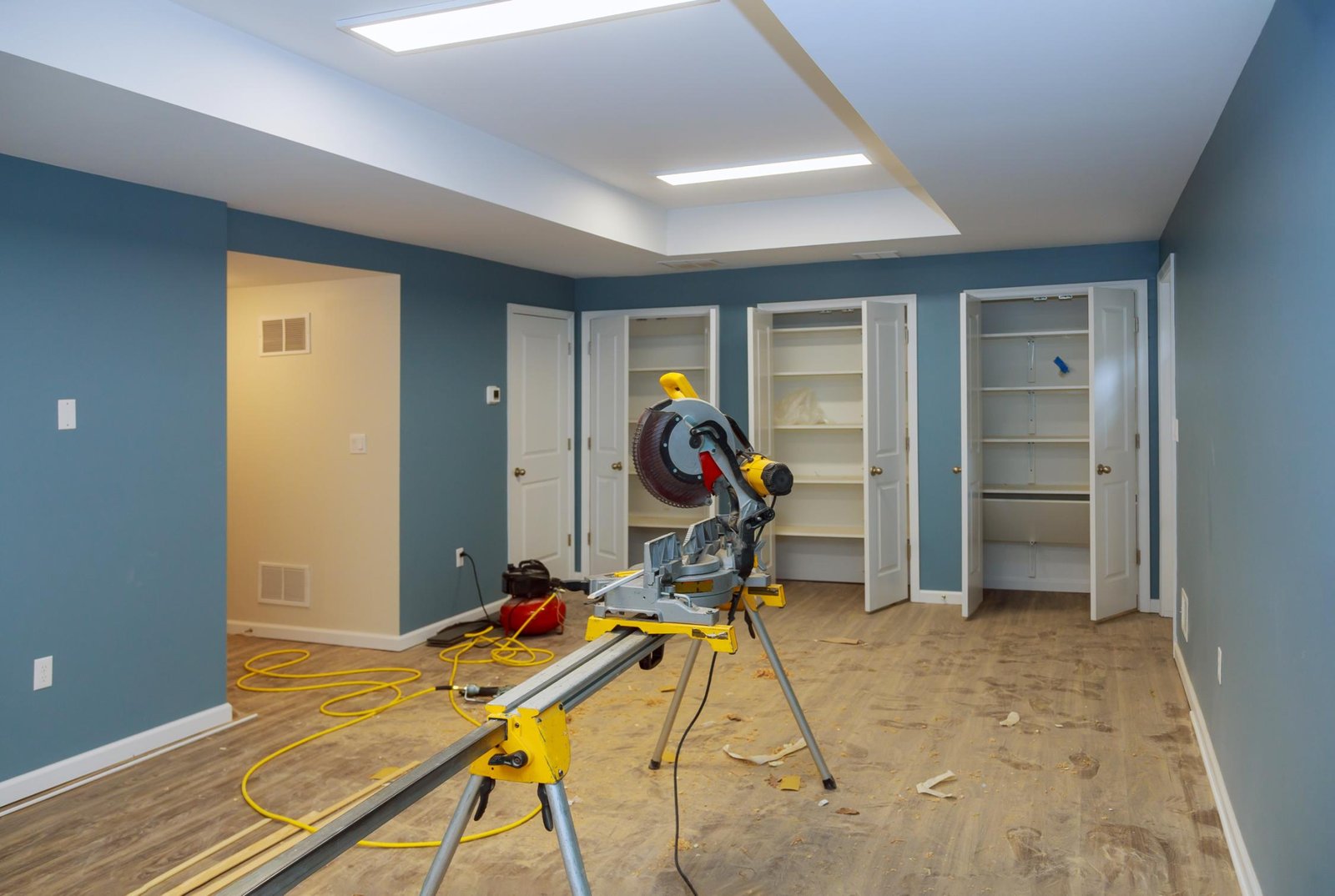 A room under renovation with blue walls, open white closets, a miter saw on a stand, wood planks, and extension cords on a wood floor.