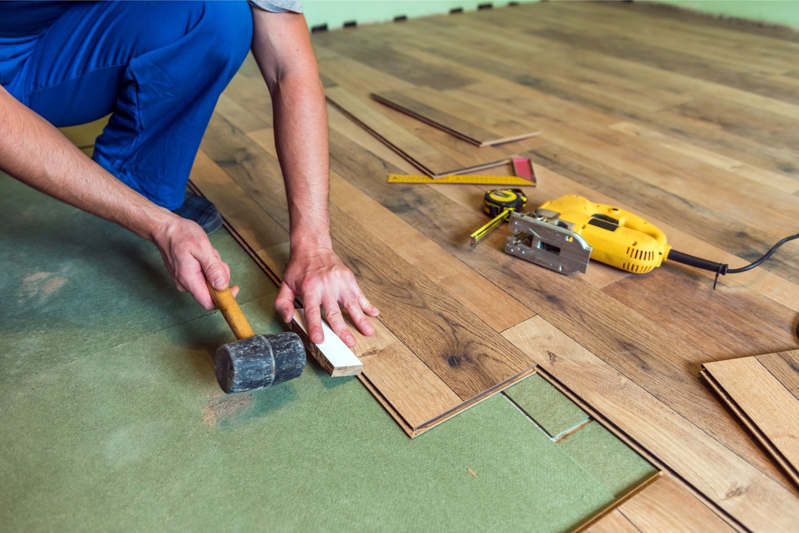 A person installs wooden laminate flooring using a mallet and spacer, with tools such as a tape measure, level, and jigsaw nearby.