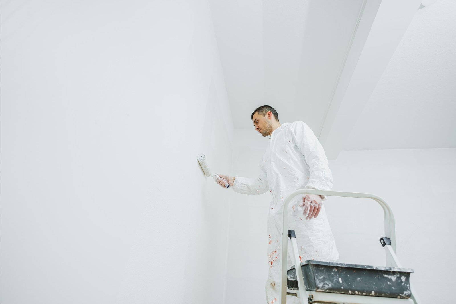 A person in a white protective suit stands on a step ladder and paints a white wall with a roller in a brightly lit room.